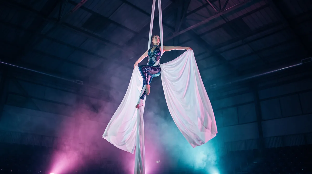 An aerialist in an extended pose on white silks, high above a darkened venue floor